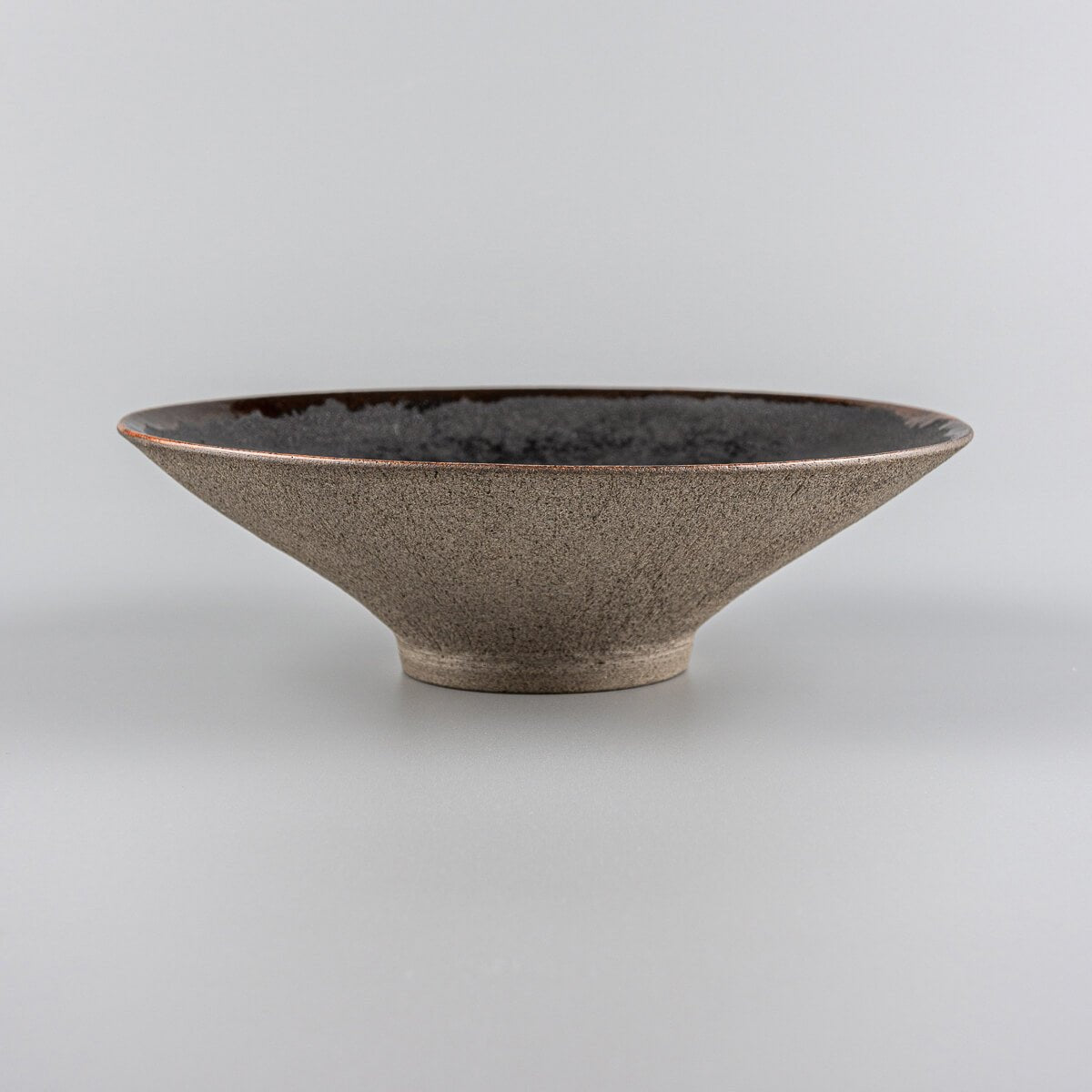 Side view of artisanal ceramic plate with elevated base, black interior, and unglazed exterior.