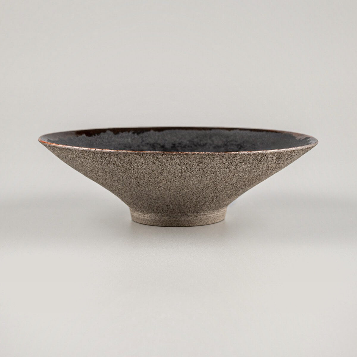 Side view of artisanal ceramic plate with elevated base, black interior, and unglazed exterior.