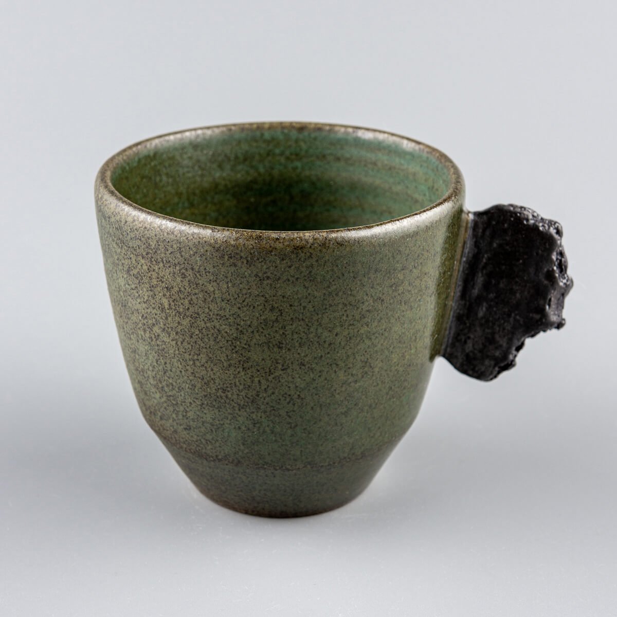 Single handmade pottery mug in dark stoneware with green glaze and sculpted black clay handle.