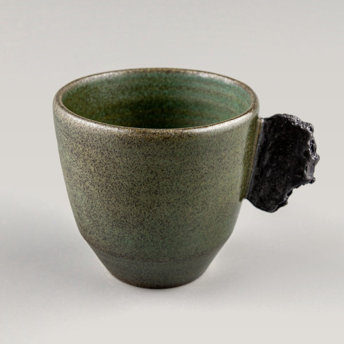 Single handmade pottery mug in dark stoneware with green glaze and sculpted black clay handle.