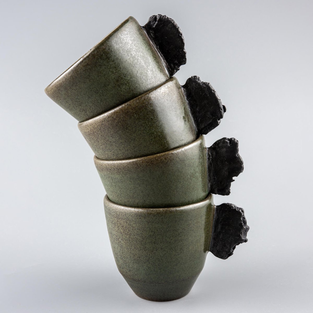 Stacked artisanal pottery mugs in matte green glaze with rustic black clay handles for modern kitchenware.