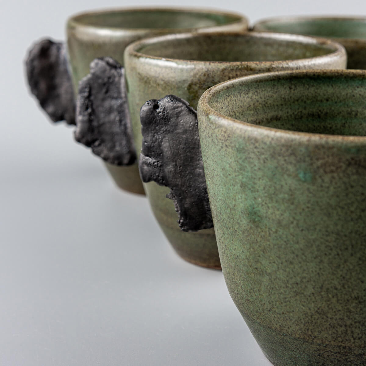 Four handcrafted pottery mugs side by side in elegant green glaze with unglazed black clay handles.