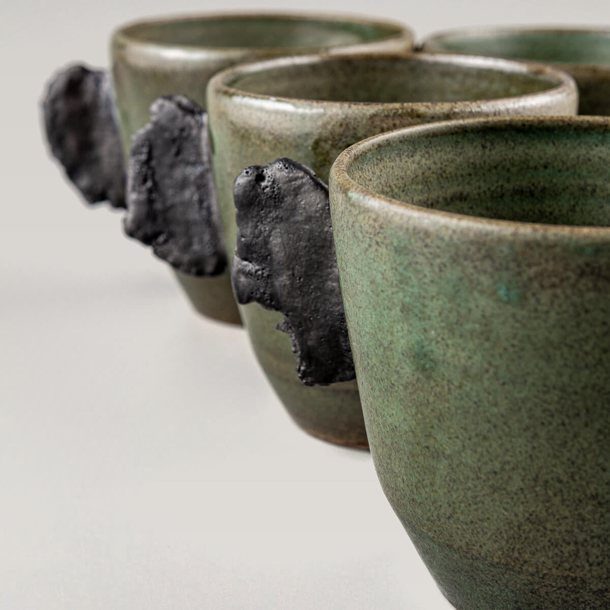 Four handcrafted pottery mugs side by side in elegant green glaze with unglazed black clay handles.