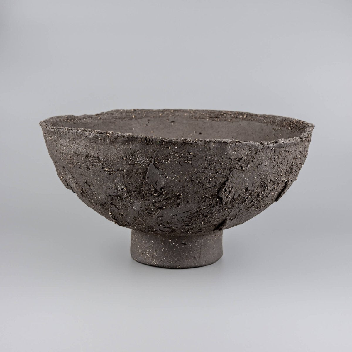Handmade decorative bowl in dark stoneware with raised foot, rough rim, and sculptural centerpiece form for interior design.