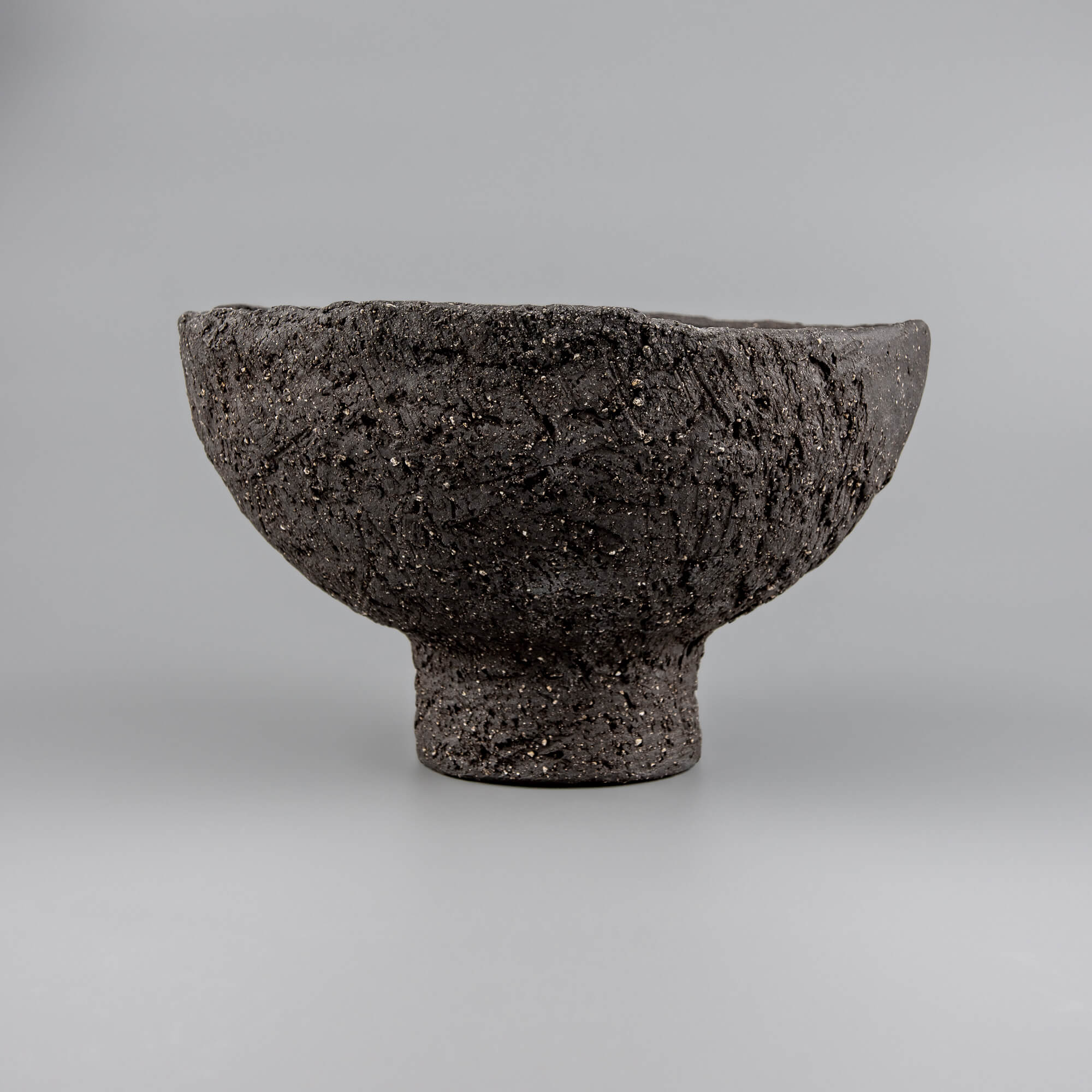 Textured handmade decorative bowl with black clay surface, pedestal base, and bold presence for home or hotel interiors.